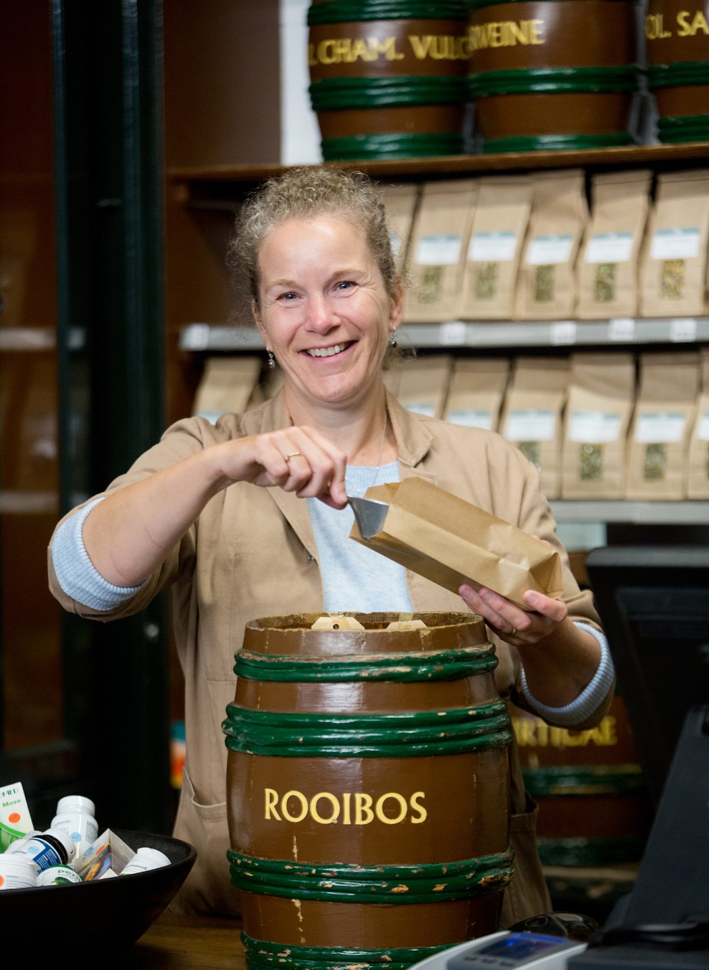 Lachende Petra van Ginkel met kruidenton met zakje en schepje in bruine stofjas
